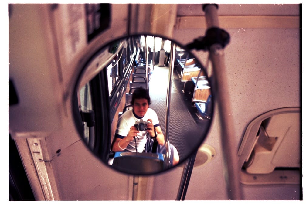 A self-portrait of Naz Hamid, taken in 2000. I'm taking a photo of myself reflected in the circular mirror of a bus.