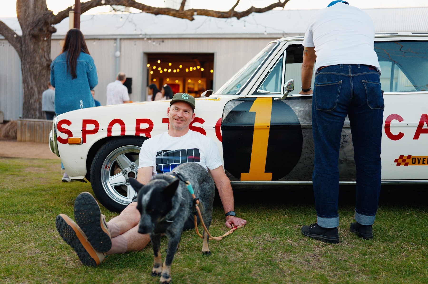 A man sits against a lovely vintage car with his dog in the grass on a sunny day.